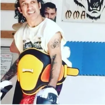 Coach Rex grinning behind his gold Muay Thai belly pad under the gym's tiger sign