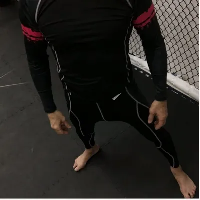 Black rash guard with red brush-stroke sleeves, modelled barefoot on the cage mat